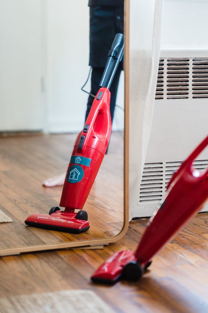A red vacuum cleaner stands on a wooden floor, reflected crisply in a mirror.